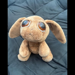 May include: A small, brown plush rabbit toy with long, floppy ears and oversized, glossy, dark amber-coloured plastic eyes. The stuffed animal has a soft, textured fabric exterior and is depicted in a seated position with its front paws resting forward.