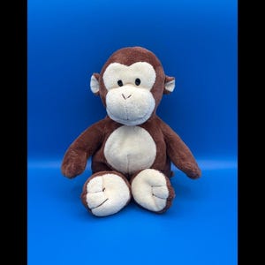 May include: A plush monkey toy featuring soft brown fur on its head, arms, and back, contrasted with a cream-coloured face, chest, and feet. The stuffed animal has small ears, a rounded snout, and stitched details on its paws, sitting upright.