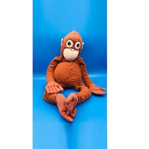 Puede incluir: Un peluche de orangután con pelaje marrón, ojos grandes y redondos y una cara blanca. El peluche está sentado con las piernas extendidas y los brazos a los lados sobre un fondo azul. El juguete mide aproximadamente 50 cm de alto.