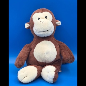 May include: A plush monkey toy featuring soft brown fur on the body, arms, and head, with a contrasting cream-colored face, belly, and feet. The monkey has small rounded ears, a stitched mouth, and dark eyes, presented in a seated position.