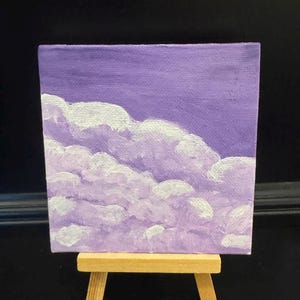 May include: A square canvas painting featuring a sky filled with fluffy white clouds against a solid purple background, displayed on a small wooden easel. The artwork uses varying shades of purple and white paint to create a textured cloud effect.