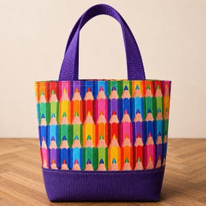 May include: A tote bag featuring a repeating pattern of colorful sharpened pencils in shades of red, orange, yellow, green, blue, and purple. The bag has a solid purple fabric base and matching purple woven fabric handles at the top.