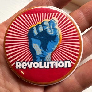 May include: A round button with a red background and white radial stripes. A blue fist is in the center, with the word "Revolution" in white text below the fist. The button has a gold border.