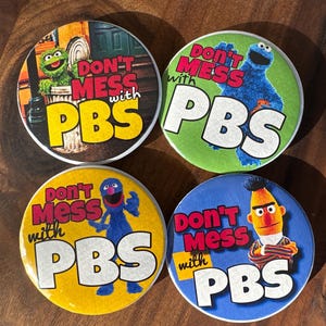 May include: Four round buttons featuring Sesame Street characters. Each button has the text "DON'T MESS with PBS". Characters include Oscar the Grouch, Cookie Monster, Grover, and Bert. The buttons are on a wooden surface.