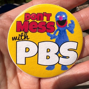 May include: A round yellow button with the text "Don't Mess with PBS" in red and white. A blue cartoon character with a pink nose and mouth is featured on the button.