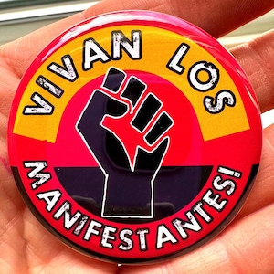May include: A round button with the text "VIVAN LOS MANIFESTANTES!" in white, arched around a black fist graphic. The background features concentric circles of red, yellow, and black.