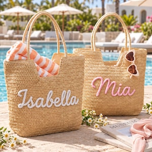May include: Two woven straw tote bags with top handles. The left bag features the white embroidered name Isabella and contains an orange and white striped towel. The right bag features the pink embroidered name Mia and heart-shaped sunglasses.