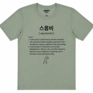 Pode incluir: Camiseta verde sálvia com o texto "스몸비 [seumombi]" e uma definição em preto. A definição descreve indivíduos absortos em seus smartphones. A camisa também apresenta uma pequena ilustração de uma pessoa olhando para um telefone.