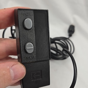 Pode incluir: Um controle remoto retangular preto da Kodak com superfície texturizada, apresentando dois botões circulares cinzas rotulados como FOR e REV. O dispositivo inclui um longo cabo preto e um plugue, com o logotipo da Kodak em relevo.