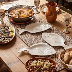 Puede incluir: Una mesa puesta con tres fuentes de cerámica blanca con forma de pescado, una olla grande de metal con guiso de marisco, una jarra de cerámica marrón, un cuenco de alubias, un plato de pescado a la parrilla y copas con motivos florales.
