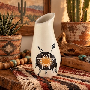 May include: A cream-colored ceramic vase with an asymmetrical slanted rim. The front features a black circular graphic containing a geometric star pattern and two crossed arrows. The vase has a smooth, matte finish and a tapered, rounded body.