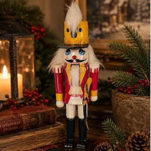 May include: A wooden nutcracker figurine wearing a yellow hat with a white plume, a red jacket with yellow accents, and black boots. The figure has a white beard, painted blue eyes, and a black mustache, holding a yellow sword at its side.