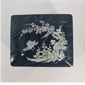 May include: A rectangular black box featuring an iridescent mother-of-pearl inlay on the lid. The design depicts two birds perched among flowering branches with delicate petals and leaves, creating a shimmering contrast against the dark surface.