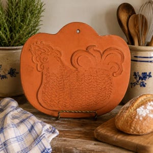 May include: A terracotta-colored ceramic plaque featuring a relief design of a rooster in profile. The plaque has a rounded, scalloped top edge with a small circular hole for hanging. The rooster is depicted with textured feathers and a large tail.