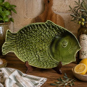 May include: A glossy, olive-green ceramic serving platter shaped like a fish, featuring a detailed embossed scale pattern across its body. The platter is supported by a dark brown wooden stand, showcasing a smooth, glazed finish and defined fins.
