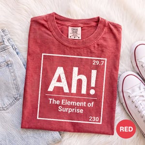 May include: A red short-sleeved t-shirt with a white graphic print resembling a periodic table element. The text reads Ah! The Element of Surprise, with the numbers 29.7 and 230. The shirt features a Comfort Colors brand label at the inner neckline.