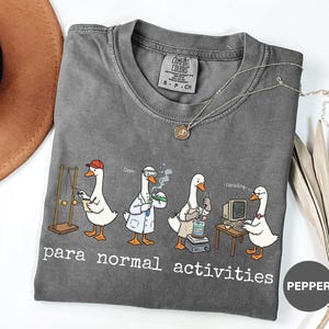 Puede incluir: Una camiseta gris Comfort Colors con una caricatura de gansos realizando actividades científicas. La frase "para normal activities" está impresa debajo. Un collar plateado con un colgante está sobre la camiseta.