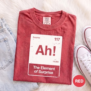 May include: A red crew neck t-shirt featuring a white rectangular graphic resembling a periodic table element. The graphic displays the text Surprise, 117, Ah!, 14.094, and The Element of Surprise. The shirt has a Comfort Colors brand label.