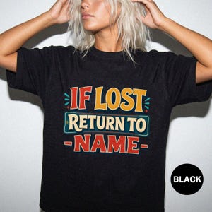 Pode incluir: Uma t-shirt preta de manga curta com uma impressão gráfica com o texto IF LOST RETURN TO NAME numa fonte de estilo retro. As letras utilizam uma paleta de cores vermelho, amarelo e creme, acentuada com pequenas marcas decorativas em azul-petróleo.