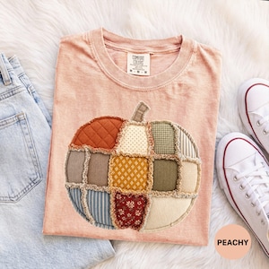 Op de afbeelding: Een perzikkleurig t-shirt met een grote, centrale pompoengrafiek gemaakt van verschillende doorgestikte stoffen patches in tinten oranje, bruin, groen en crème. De pompoen heeft een applicatiestijl met gerafelde randen. Het t-shirt heeft een ronde hals en een Comfort Colors-label.
