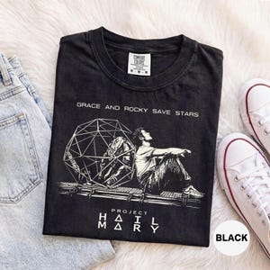 Può includere: Una t-shirt nera a maniche corte con una grafica bianca che raffigura una persona seduta accanto a una struttura sferica geometrica. Il testo recita "GRACE AND ROCKY SAVE STARS" e "PROJECT HAIL MARY". La maglietta ha un girocollo e un'etichetta taglia M.