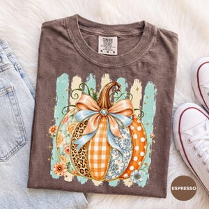 Op de afbeelding: Een bruin t-shirt met een afbeelding van een pompoen versierd met een grote lichtblauwe strik. Het pompoenontwerp bevat secties met luipaardprint, oranje gingham, bloemmotieven en oranje stippen, tegen een lichtblauwe gestreepte achtergrond.
