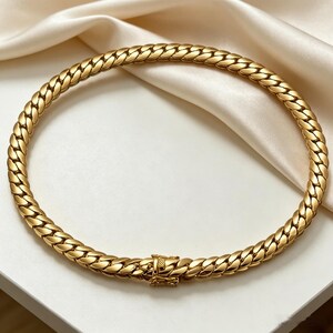K&ouml;nnte beinhalten: Eine goldfarbene Halskette mit einem geflochtenen Kettenmuster. Die Halskette ist kreisf&ouml;rmig und hat einen Verschluss. Der Schmuck wird auf einer wei&szlig;en Oberfl&auml;che mit einem cremefarbenen Satinstoff im Hintergrund pr&auml;sentiert.