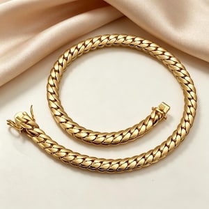 K&ouml;nnte beinhalten: Eine goldfarbene Halskette mit einem Curb-Chain-Design. Die Halskette besteht aus Metall und verf&uuml;gt &uuml;ber einen sicheren Verschluss. Die Kette ist dick und hat ein gewebtes Aussehen. Die Halskette wird auf einer wei&szlig;en Oberfl&auml;che mit einem pfirsichfarbenen Stoff im Hintergrund pr&auml;sentiert.