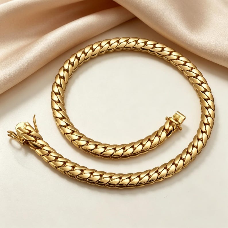 K&ouml;nnte beinhalten: Eine goldfarbene Halskette mit einem Curb-Chain-Design. Die Halskette besteht aus Metall und verf&uuml;gt &uuml;ber einen sicheren Verschluss. Die Kette ist dick und hat ein gewebtes Aussehen. Die Halskette wird auf einer wei&szlig;en Oberfl&auml;che mit einem pfirsichfarbenen Stoff im Hintergrund pr&auml;sentiert.