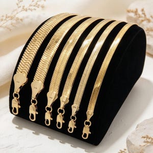 May include: Six gold-toned herringbone chains of varying widths displayed on a black velvet stand. Each chain features a flat, interlocking link pattern and a lobster claw clasp. The end tags on each chain are clearly stamped with the text 14K.
