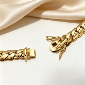 K&ouml;nnte beinhalten: Nahaufnahme eines goldenen Kettenarmbands mit Verschluss. Das Armband besteht aus dicken, ineinandergreifenden Gliedern. Der Verschluss ist ge&ouml;ffnet und zeigt den komplizierten Mechanismus. Der Hintergrund zeigt eine wei&szlig;e Oberfl&auml;che und einen drapierten, cremefarbenen Stoff.