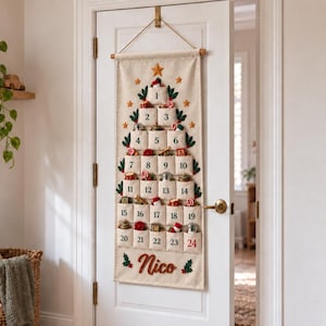 Op de afbeelding: Een cr&egrave;mekleurige stoffen adventskalender die aan een deur hangt, in de vorm van een kerstboom met vierentwintig genummerde vakjes. Elk vakje bevat kleine feestelijke voorwerpen. De naam Nico is onderaan in het rood geborduurd met hulstblaadjes.