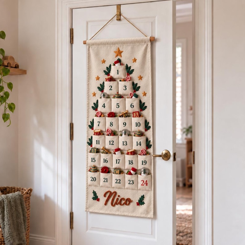 Op de afbeelding: Een cr&egrave;mekleurige stoffen adventskalender die aan een deur hangt, in de vorm van een kerstboom met vierentwintig genummerde vakjes. Elk vakje bevat kleine feestelijke voorwerpen. De naam Nico is onderaan in het rood geborduurd met hulstblaadjes.