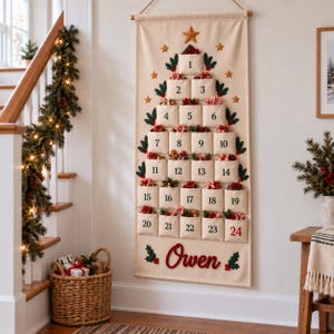 Op de afbeelding: Een cr&egrave;mekleurige stoffen adventskalender voor aan de muur in de vorm van een kerstboom met vierentwintig genummerde vakjes. Elk vakje is voorzien van groene hulstblaadjes en rode bessen. De naam Owen is onderaan in rode letters geborduurd.