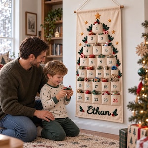Op de afbeelding: Een cr&egrave;mekleurige stoffen adventskalender voor aan de muur in de vorm van een kerstboom met vierentwintig genummerde vakjes. De kalender is voorzien van geborduurde groene hulstblaadjes, gouden sterren en de naam Ethan in donkergroen.