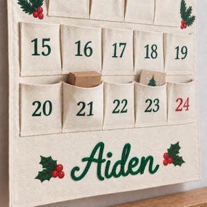 Op de afbeelding: Een cr&egrave;mekleurige stoffen adventskalender met rijen zakjes genummerd van 15 tot 24 in donkergroen, waarbij het getal 24 in het rood is. De naam Aiden is onderaan in donkergroen cursief geborduurd, geflankeerd door hulst- en bessenmotieven.