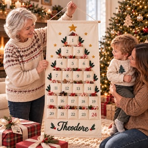 Op de afbeelding: Een cr&egrave;mekleurige stoffen adventskalender die verticaal hangt, met een opgedrukte kerstboomvorm en vierentwintig genummerde vakjes. Elk vakje is versierd met kleine feestelijke accenten. De naam Theodore staat onderaan in donkergroen cursief schrift.