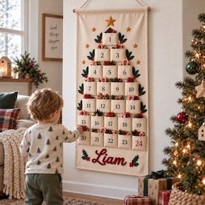 Op de afbeelding: Een cr&egrave;mekleurige stoffen adventskalender voor aan de muur in de vorm van een kerstboom met vierentwintig genummerde vakjes. De kalender is voorzien van groene bladeren, gouden sterren en de naam Liam in rode letters onderaan.