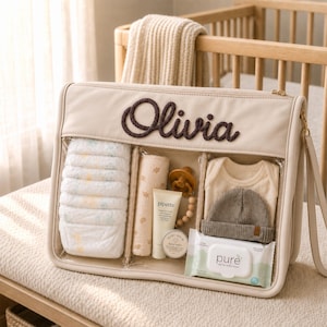 Könnte beinhalten: Eine beigefarbene, rechteckige Organisationstasche mit dem Namen Olivia, der in dunkelbrauner Seiloptik aufgestickt ist. Die Tasche hat transparente Frontpaneele, die einen Stapel Windeln, ein gerolltes Tuch, eine Pipette-Creme, einen Schnuller, einen Body, eine Mütze und Feuchttücher zeigen.
