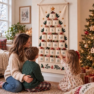 Op de afbeelding: Een cr&egrave;mekleurige stoffen adventskalender voor aan de muur in de vorm van een kerstboom met vierentwintig genummerde vakjes. De kalender is voorzien van geborduurde hulstblaadjes, sterren en de gepersonaliseerde rode en groene tekst Leo & Mia.
