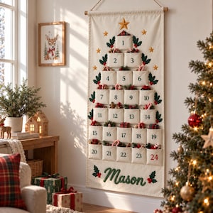 Op de afbeelding: Een cr&egrave;mekleurige stoffen adventskalender voor aan de muur in de vorm van een kerstboom met vierentwintig genummerde vakjes. De vakjes bevatten kleine feestelijke items en de naam Mason is onderaan in het groen geborduurd met hulst.