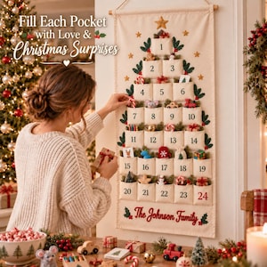 Op de afbeelding: Een cr&egrave;mekleurige stoffen wandhanger in de vorm van een kerstboom met vierentwintig genummerde vakjes. De tekst luidt Fill Each Pocket with Love & Christmas Surprises en The Johnson Family. Het heeft groene bladeren en gouden sterren als accenten.
