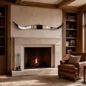 May include: A long, curved set of steer horns mounted horizontally on a wall. The horns feature a gradient of white and grey tones, transitioning to dark black tips, with a central dark brown leather-wrapped section and a small metal hanging loop.