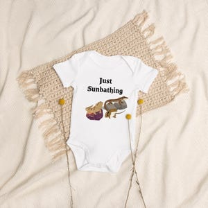 May include: White baby bodysuit with the text "Just Sunbathing" and a graphic of two lizards sunbathing on rocks. The bodysuit is made of soft material and is perfect for a baby.