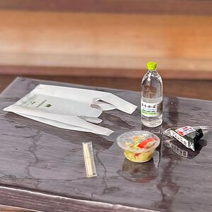 以下が含まれることがあります： 緑色のキャップが付いた透明なプラスチック製の水ボトル、白いプラスチック製の買い物袋、赤と緑の具材が見える小さな丸いサラダ容器、包装された箸、三角形のおにぎりが含まれるミニチュアセット。