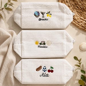 May include: Three white rectangular zippered pouches with a textured waffle-weave fabric. Each pouch features custom embroidery: the first shows a cupcake, martini, and lemons with the name Brooke; the second, a sun, golf cart, and shell with the name Maddie; the third, a hot dog, bikini, and cherries with the name Ala.
