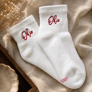 May include: A pair of white ankle socks featuring the word Ohio embroidered in red cursive script on the ribbed cuff. The socks have a soft texture and a clean design, with the word Ohio clearly visible on the upper portion of each sock.