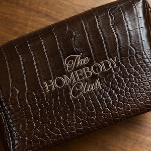 May include: A rectangular dark brown pouch with a glossy crocodile-embossed texture. The front features the text The HOMEBODY Club embroidered in a light gold thread. The item has a structured shape with visible stitching along the edges.