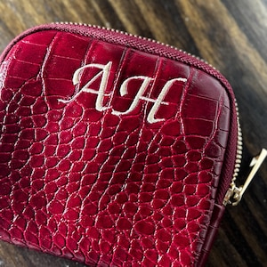 May include: A deep red, square-shaped zippered pouch featuring a textured crocodile-skin pattern. The front of the pouch is embroidered with the white monogram letters AJH in a cursive script. The pouch has a matching red zipper closure.