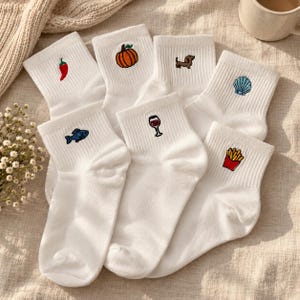 May include: A collection of white ankle socks, each featuring a small embroidered icon including a red chili pepper, an orange pumpkin, a brown dachshund, a blue seashell, a blue fish, a glass of red wine, and a red carton of french fries.